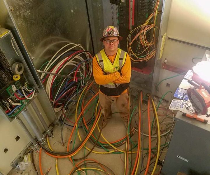A professional electrician stands amidst a complex array of colorful electrical wires and circuitry inside an industrial electrical panel room.